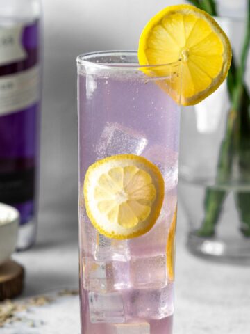 A purple gin collins in a skinny glass garnished with lemon wheels and a bottle of gin and flowers behind it.