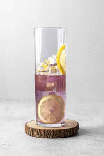 A tall skinny glass with ice, lemon wheels and a purple gin cocktail on a wooden coaster.
