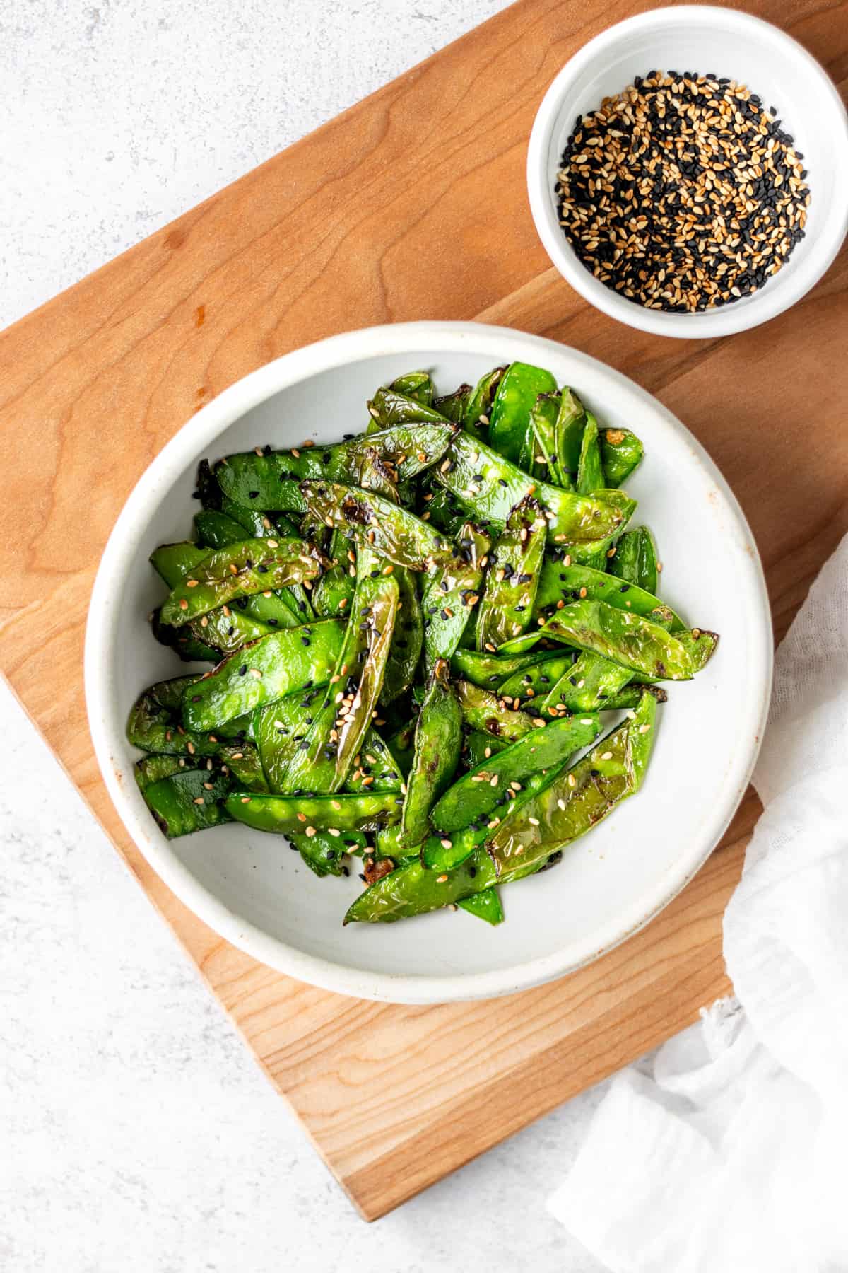 Air fried snow peas in a while bowl on a a wooden cutting board garnished with toasted sesame seeds which are also in a small white bowl.