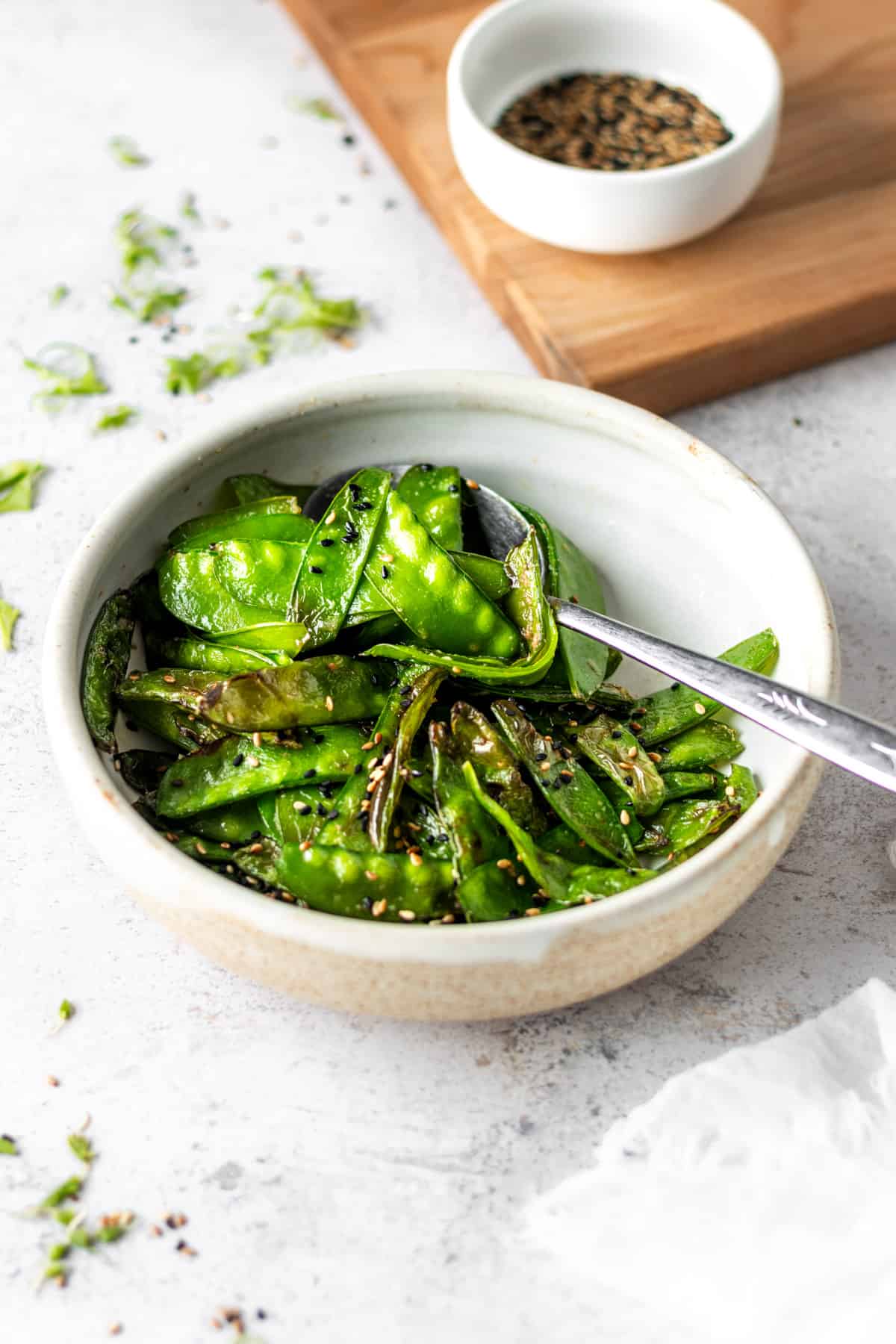 A white bowl of air fried snow peas with a spoon, garnished with sesame seeds.