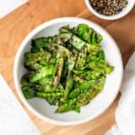 A white bowl of air fried snow peas on a wooden cutting board sprinkled with toasted sesame seeds.