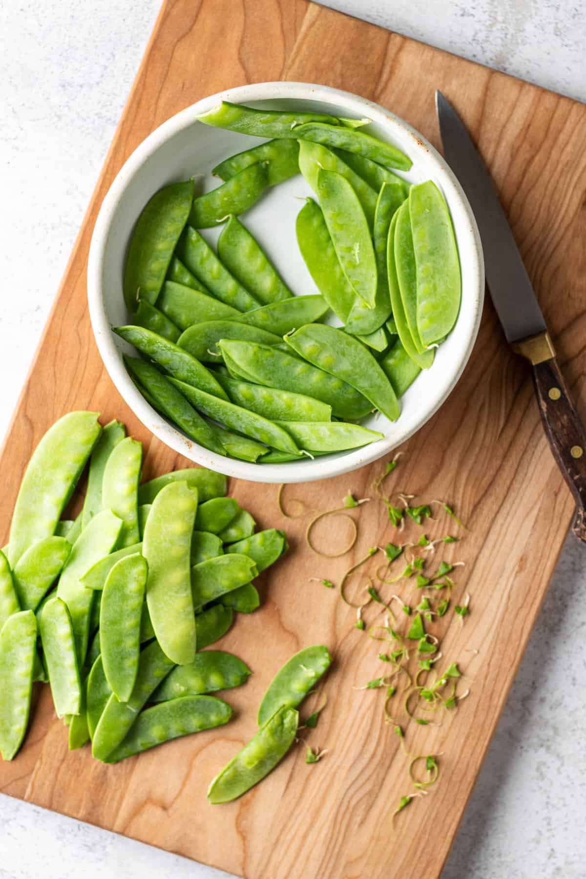 Trimmed snow peas on a wooden cutting board with a knife and bowl.