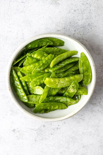 A white bowl of snow peas tossed in olive oil and spices.