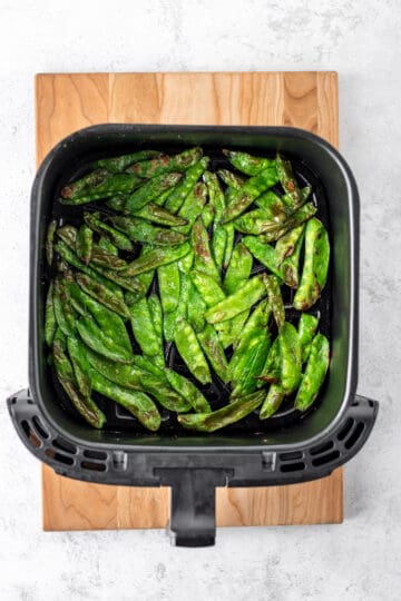 A black air fryer basked on a wooden cutting board with cooked snow peas.