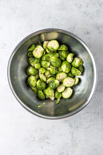 A large metal mixing bowl with halved Brussels sprouts tossed in olive oil and spices.