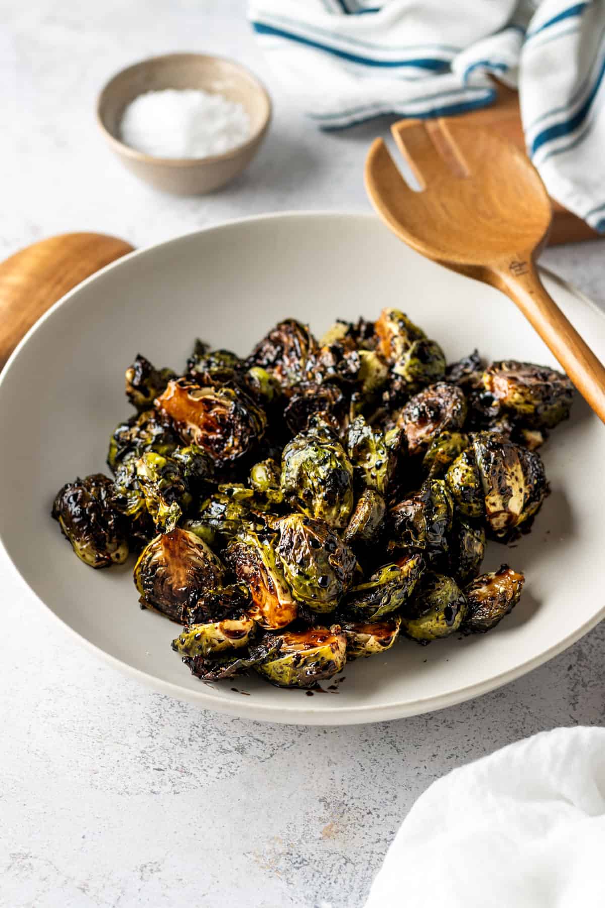 A while bowl filled with Roasted Brussels sprouts and balsamic glaze with large wooden serving utensils.