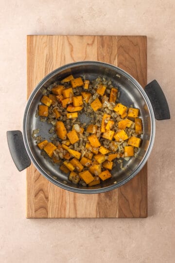 A large pot on a wooden cutting board with sautéed butternut squash, onions and spices.