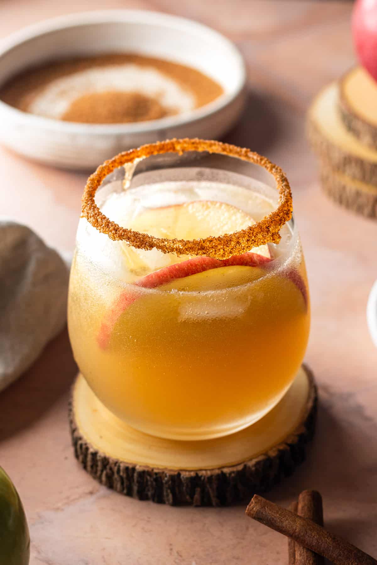A fall margarita on a wooden coaster in a wine glass garnished with cinnamon sugar and apple slices with a bowl of cinnamon sugar in the background.