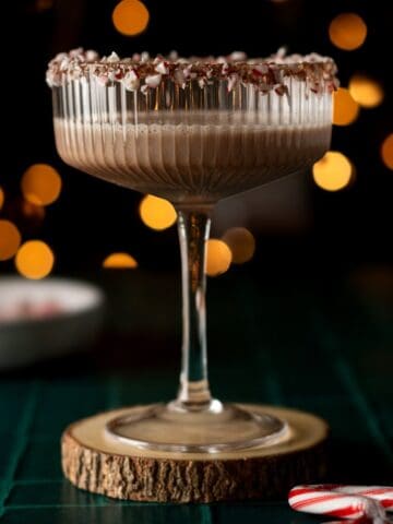 Straight on view of a coupe glasses with a candy cane rim filled with a chocolate peppermint martini with blurred lights in the background.