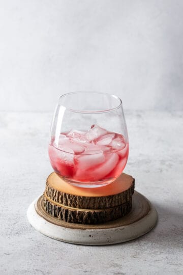 A stemless wine glass with cranberry juice and ice standing on two wooden coasters and a white plate.