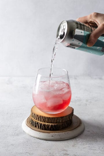 Club soda from a can being poured into a stemless wine glass filled with ice, cranberry juice, gin and simple syrup.