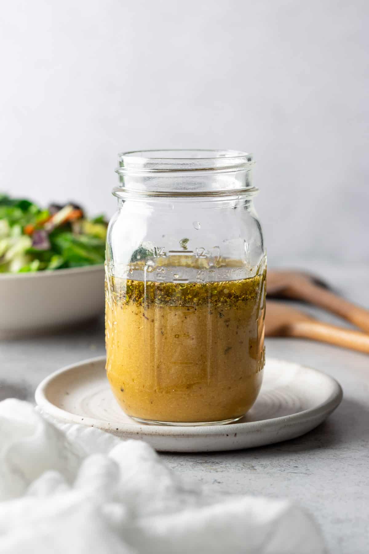 A mason jar with nutritional yeast salad dressing on a small white plate with a salad in the background.