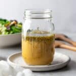 A mason jar filled with salad dressing on a small white plate with a salad in the background.