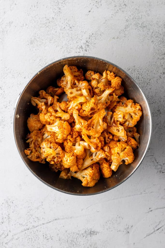 A mixing bowl with roasted cauliflower florets tossed in buffalo sauce.