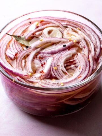 A glass bowl with pickled red onions in a brine with a bayleaf, crushed red pepper and black pepper.