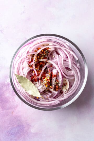Overhead of a pyrex bowl with thinly sliced red onions, a bayleaf, black pepper and crushed red pepper.