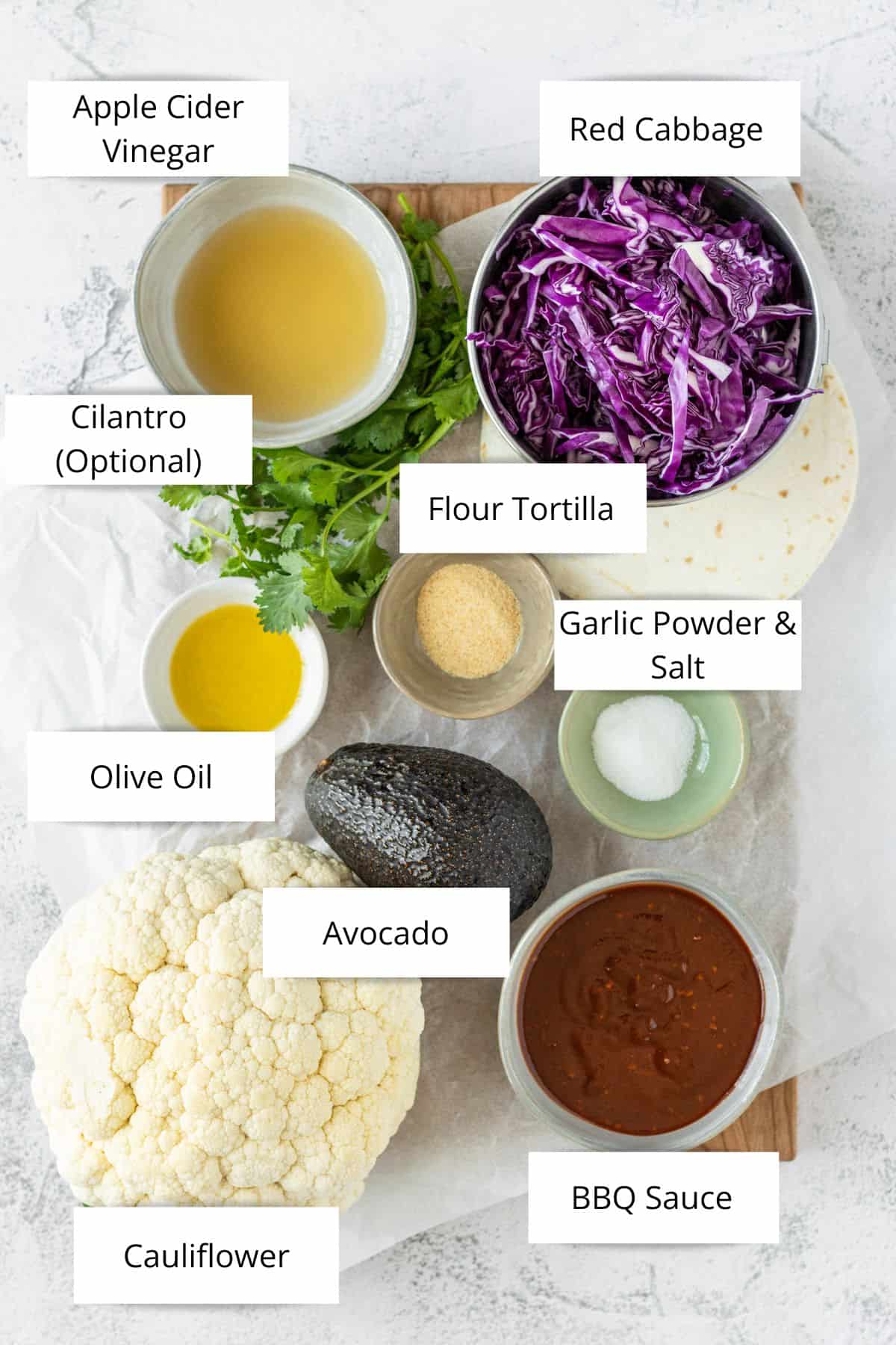 Overhead of a bowl of apple cider vinegar, sliced red cabbage, flour tortillas, pinch bowls of spices and oil, an avocado, a head of cauliflower and a bowl of barbecue sauce.
