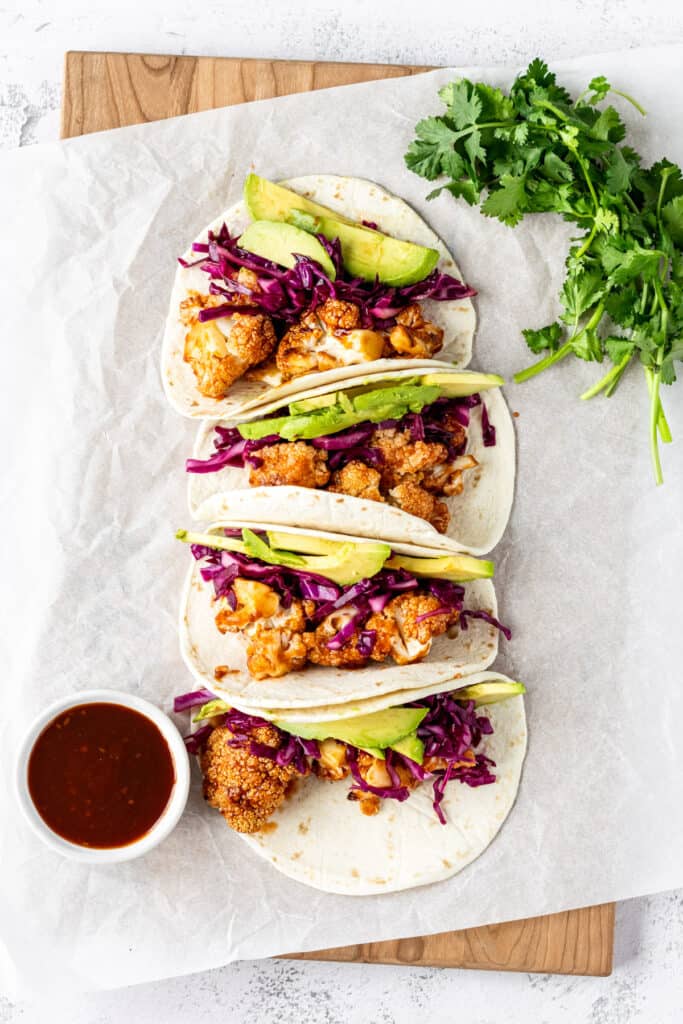 Overhead of 4 cauliflower tacos on a wooden cutting board with a small bowl of bbq sauce to the side.
