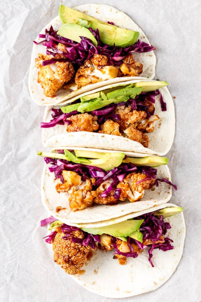 Overhead view of 4 cauliflower tacos with red cabbage and avocados in soft tortillas on a white background.