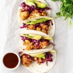 Overhead view of cauliflower tacos with red cabbage and avocados in soft tortillas on a white background with a bowl of bbq sauce.