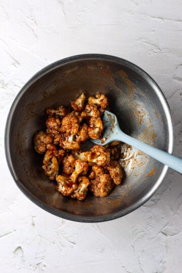 A large mixing bowl with cauliflower tossed in bbq sauce with a blue spatula.