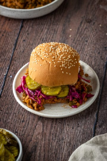 A sesame burger bun with lentil sloppy Joe, red cabbage and pickles on a small white plate on a wooden background.
