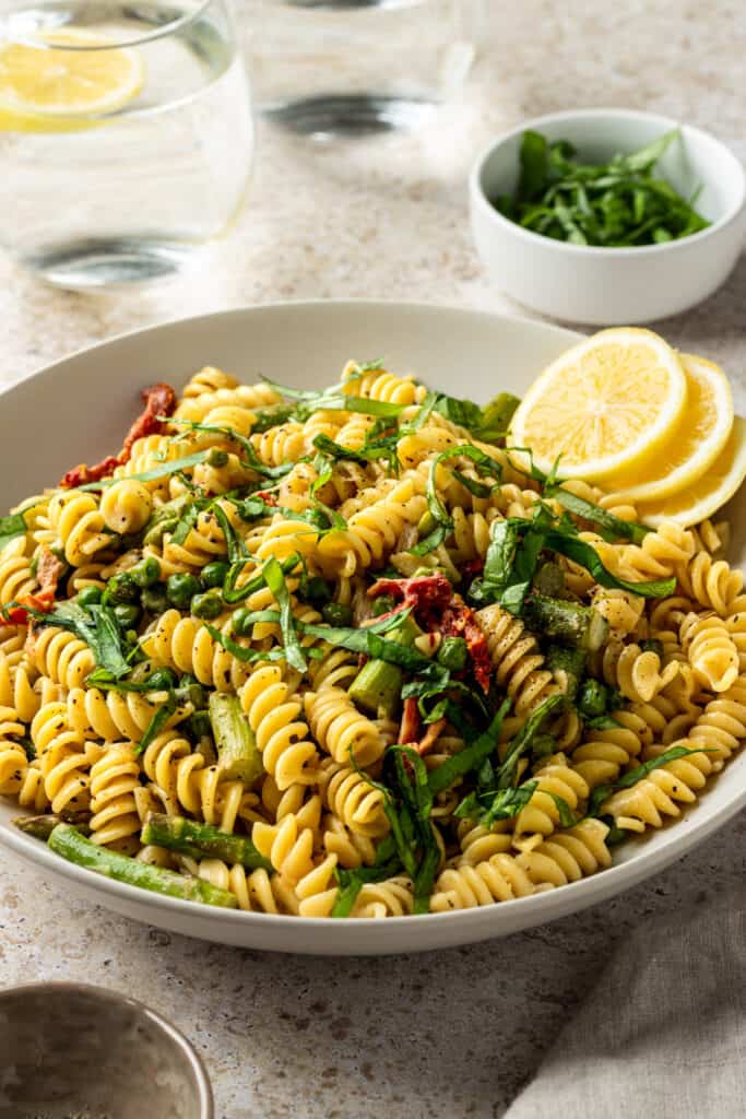 A bowl of asparagus pea pasta with sun-dried tomatoes garnished with lemon wheels and basil.