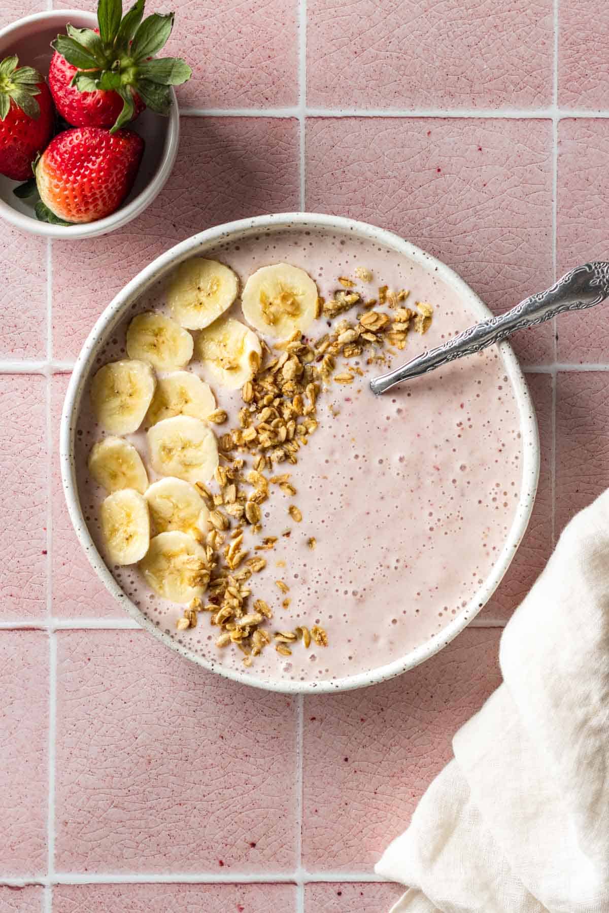 A strawberry banana smoothie bowl with a spoon garnished with granola and banana slices.