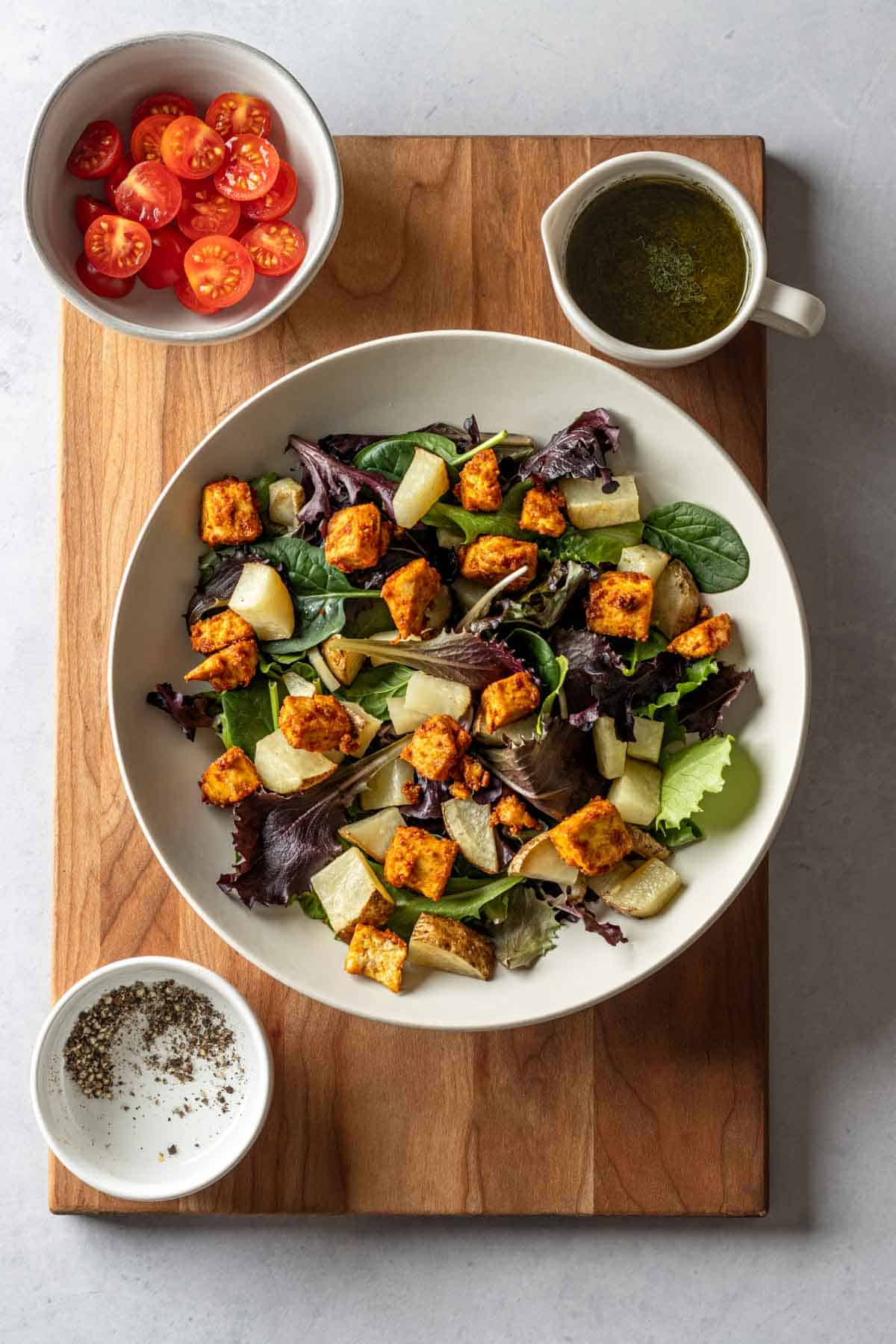 A wooden cutting board with a bowl of salad with toppings on the side.