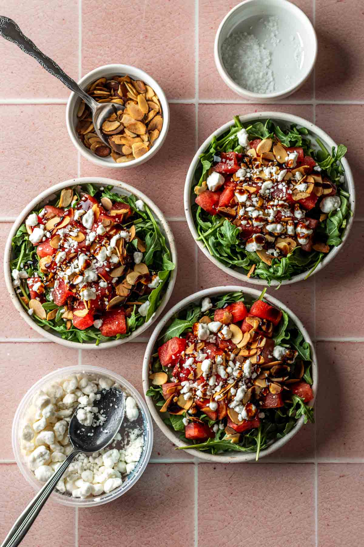 There bowls of watermelon arugula salad with side dish of sliced almonds and goat cheese.