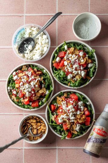 Three bowls of watermelon arugula salad with sides of cheese and almonds.