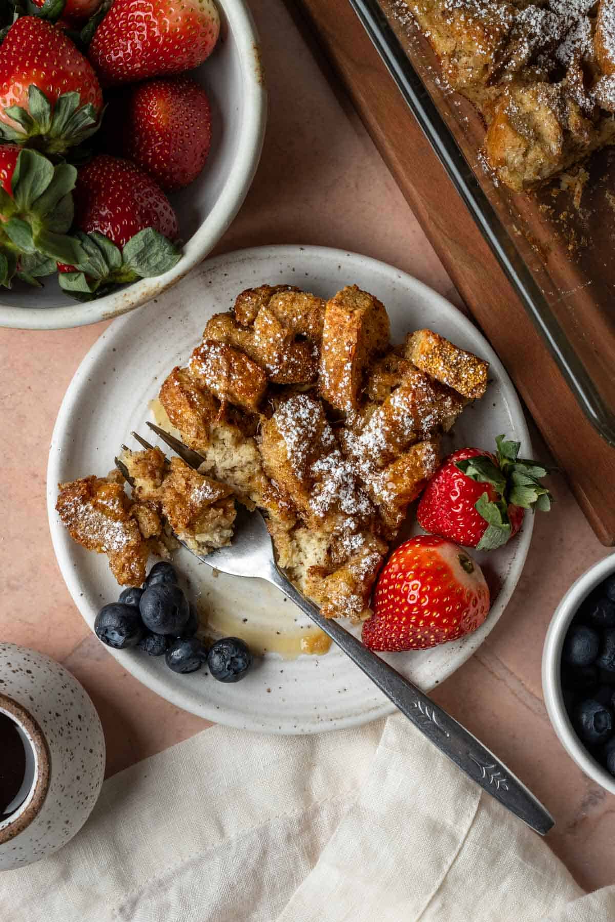 A slice of French toast casserole on a plate with strawberries, blueberries and maple syrup.