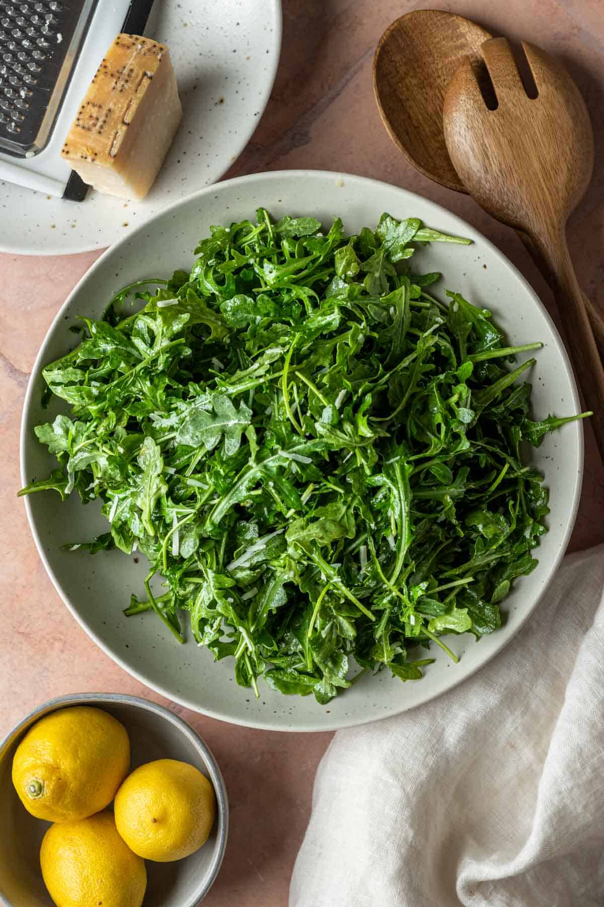Arugula parmesan salad in a bowl with lemons on the side and a block of cheese.